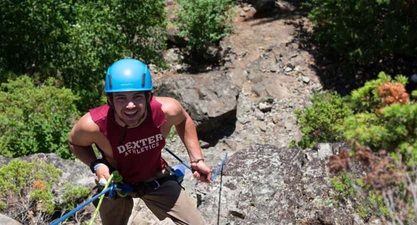 A person wearing safety gear is secured by ropes as they look up at the camera and smile while rock climbing.